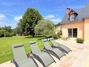 Eine Terrasse mit vier Liegestühlen, ein Haus mit Satteldach und ein großer Baum im Hintergrund.