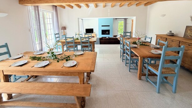 Un comedor con mesas de madera y sillas azules, una cocina al fondo y una sala de estar con televisor.