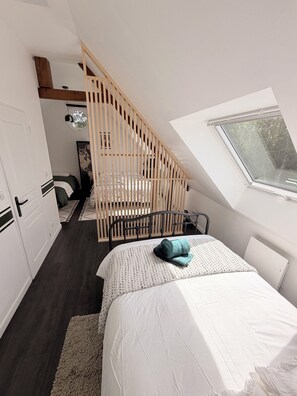 Ein Schlafzimmer mit schrägverdeckter Decke, einem Bett mit weißer und gemusterter Bettdecke, einem Holz-Kopfschild, einem Fenster mit Ausblick und einer Treppe mit Holzwand.