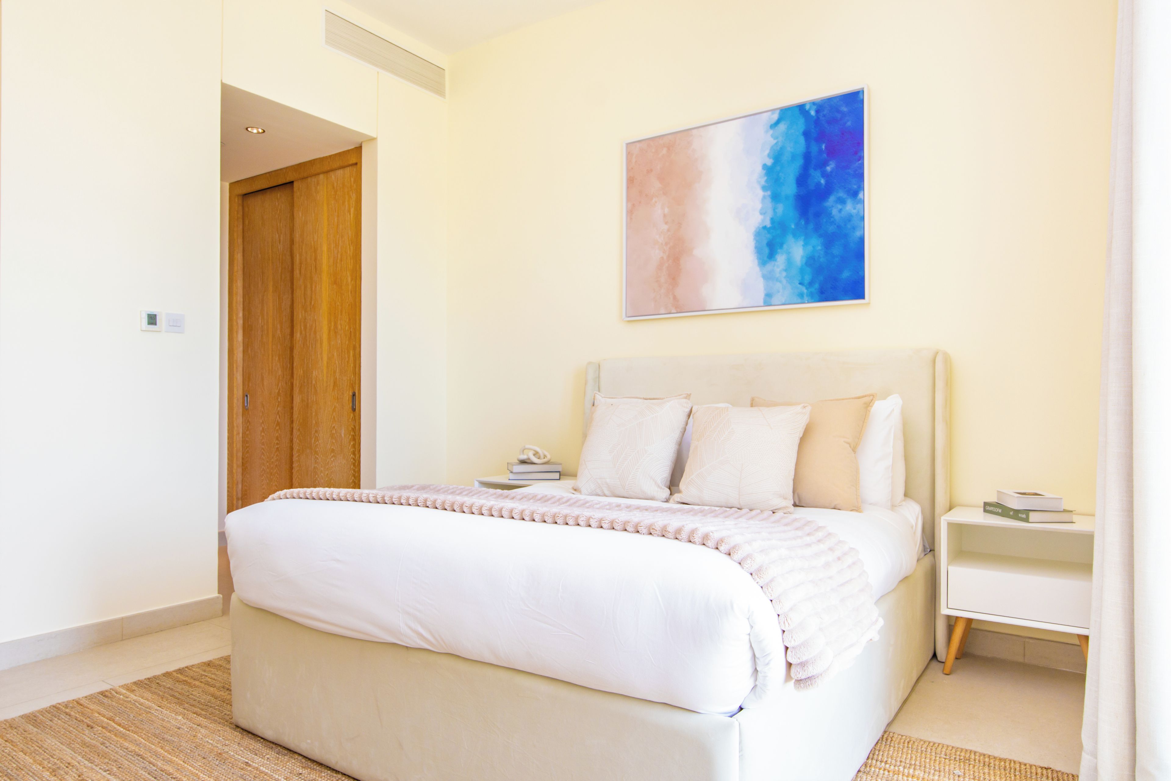 A bedroom with a bed, bedside tables, a nightstand, a wooden door, and a wall-mounted painting.