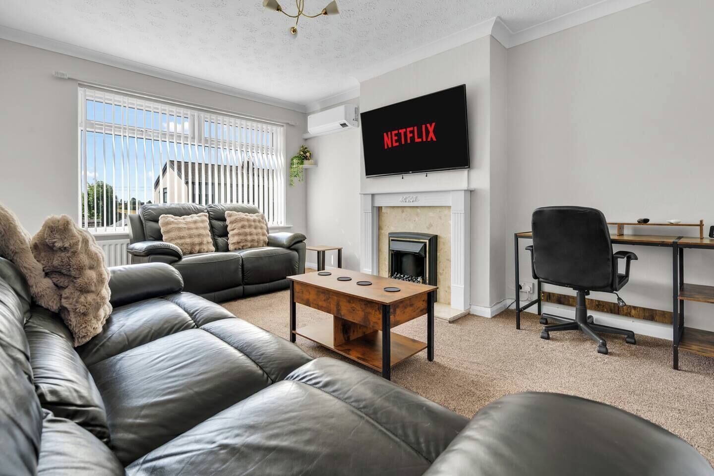 A living room with a leather sofa, a coffee table, a fireplace, a TV mounted on the wall, and a desk with a chair.