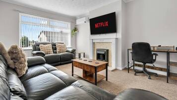 A living room with a leather sofa, a coffee table, a fireplace, a TV mounted on the wall, and a desk with a chair.