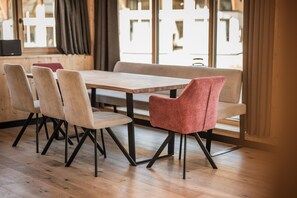 Un comedor con una mesa de madera y cuatro sillas, una de ellas con cojín rojo, en una habitación con ventanas amplias y piso de madera.