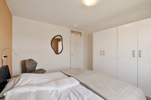 Ein Hotelzimmer mit Bett, Sessel, Schreibtisch mit Lampe, Kleiderschrank und Spiegel an der Wand.