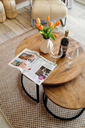Ein runder Holztisch mit einer weißen Vase, in der orangefarbene Tulpen stehen, einer Weinflasche und zwei Weingläsern. Auf dem Tisch liegt eine aufgeschlagene Zeitschrift.