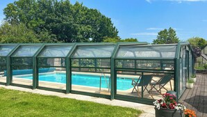 Ein Schwimmbecken mit Glasüberdachung, umgeben von einer Rasenfläche und Topfpflanzen.