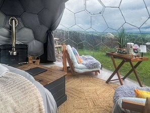 Una habitación con un muro de vidrio hexagonal, un sillón acogedor con un cojín mullido, una mesa de madera y una mesita auxiliar con una lámpara.