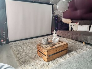 Una habitación con pantalla de proyector, un cofre de madera con objetos decorativos, un sofá con cojines y un altavoz.