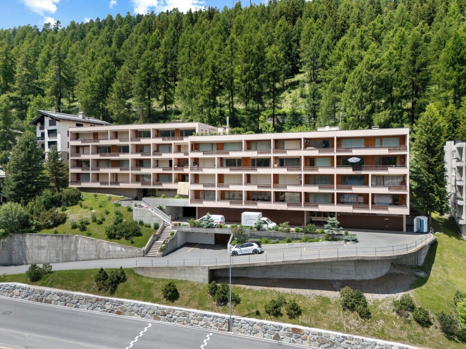 Ein mehrstöckiges Hotel mit Balkonen, einem Parkplatz und einem bewaldeten Hügel im Hintergrund.
