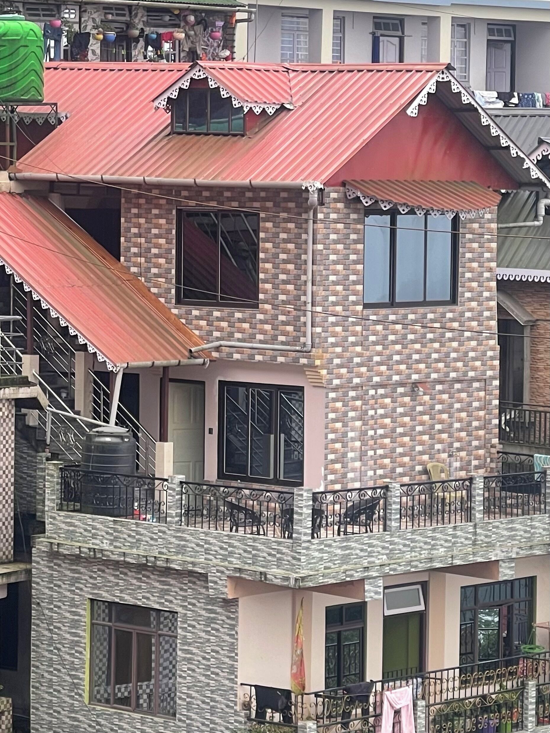 Un bâtiment à deux étages, avec un toit rouge, un balcon doté de rampes en métal et une fenêtre ornée d’un rideau.