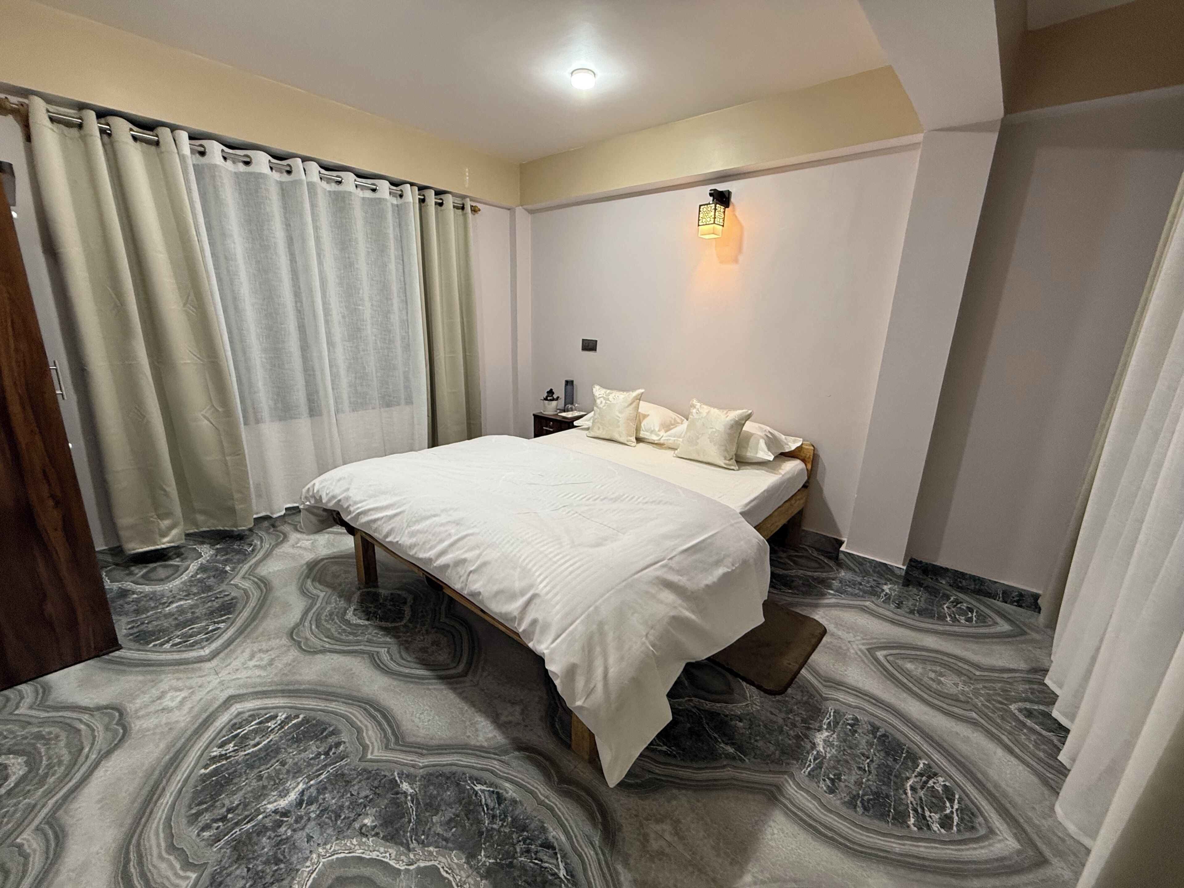 Une chambre à coucher avec un lit, des rideaux, une table de chevet, une lampe fixée au mur et un sol à motifs de marbre.