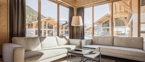 Un interior moderno con un sofá, una mesa de centro y ventanas amplias que ofrecen vistas a casas de montaña.