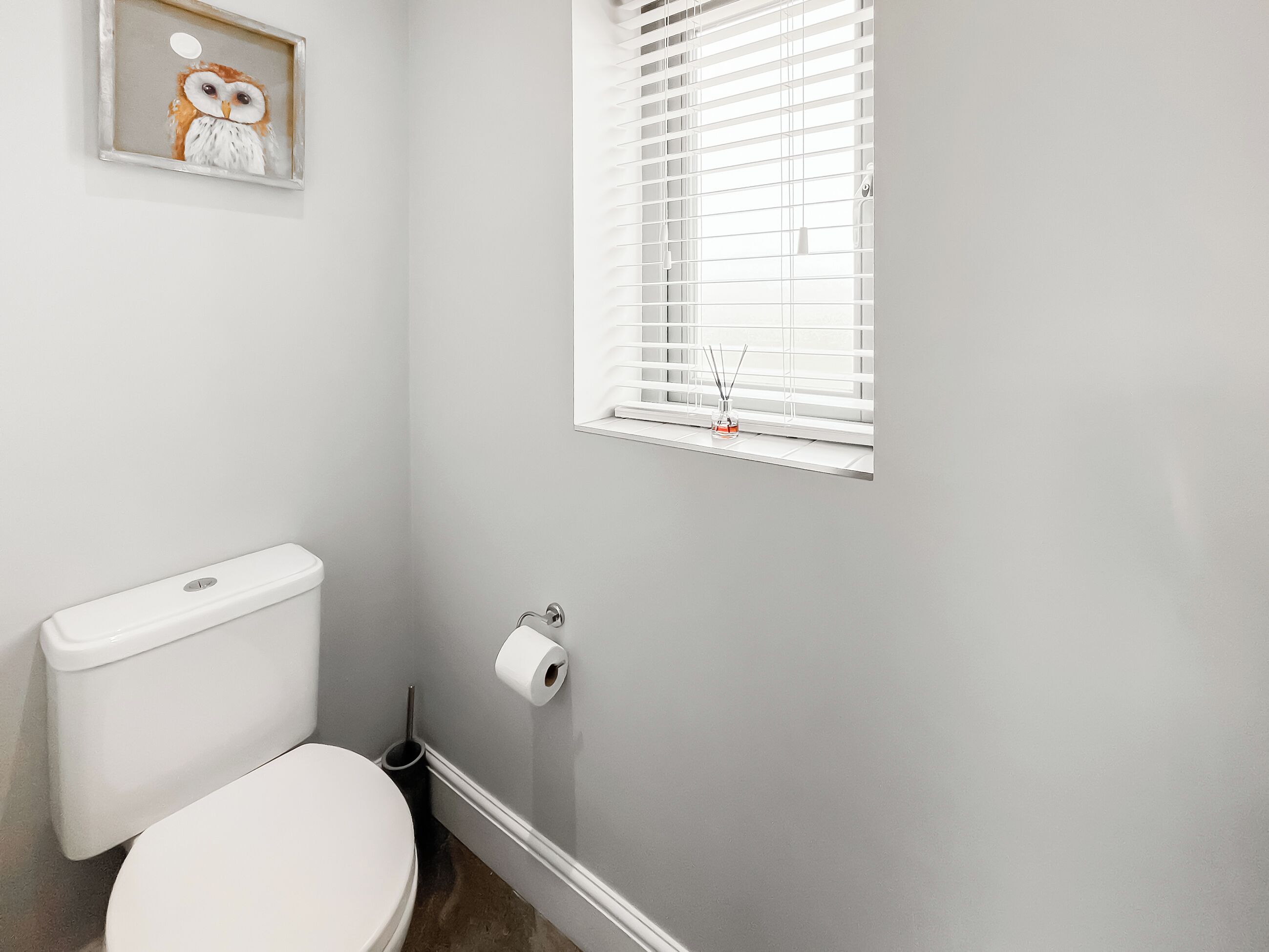 Un baño con inodoro, un rollo de papel higiénico, una ventana con persianas y un marco de fotos en la pared.