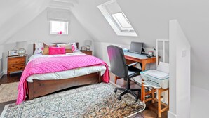 Ein Schlafzimmer mit schrägverdeckter Decke, einem Oberlicht, einem Bett mit rosa Quilzdecke, einem Schreibtisch mit Computer und einem gemusterten Teppich.