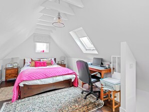 Ein Schlafzimmer mit schrägverdeckter Decke, einem Oberlicht, einem Bett mit rosa Quilzdecke, einem Schreibtisch mit Computer und einem gemusterten Teppich.