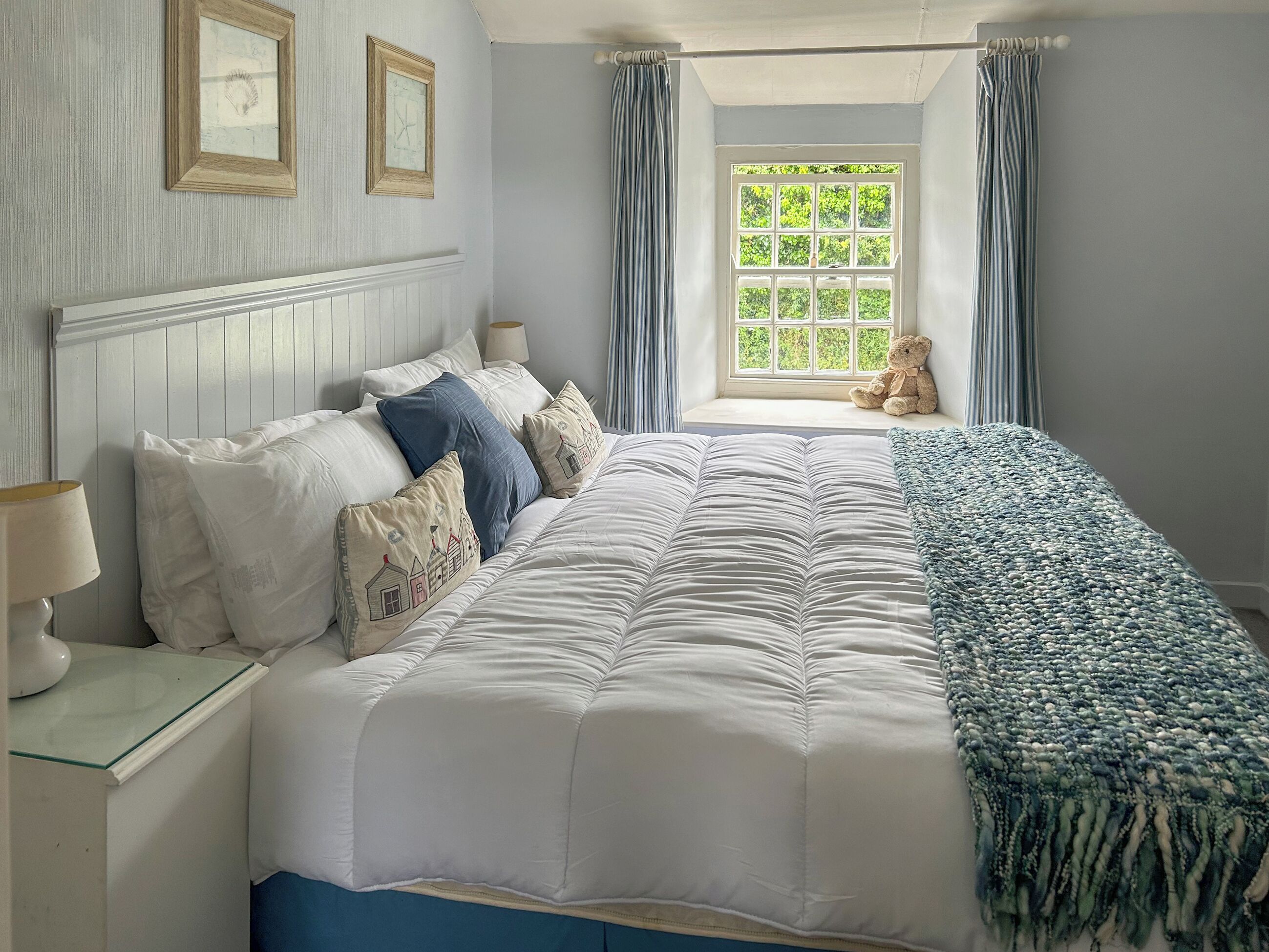 A bedroom with a large bed, a window with curtains, a teddy bear, and two framed pictures on the wall.