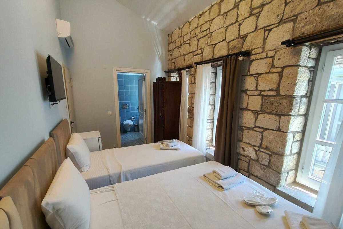 Habitación de hotel con dos camas, una pared de piedra, un televisor y un baño visible a través de una puerta abierta.