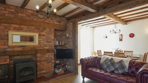Una sala de estar con vigas de madera a la vista, una chimenea de ladrillo, un sofá de cuero y un comedor con candelabro.
