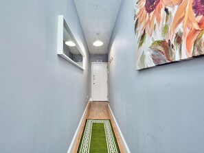 Ein schmaler Flur mit einer weißen Tür am Ende, einem Teppich mit grün-weißem Muster und einem großen floralen Gemälde an der Wand.
