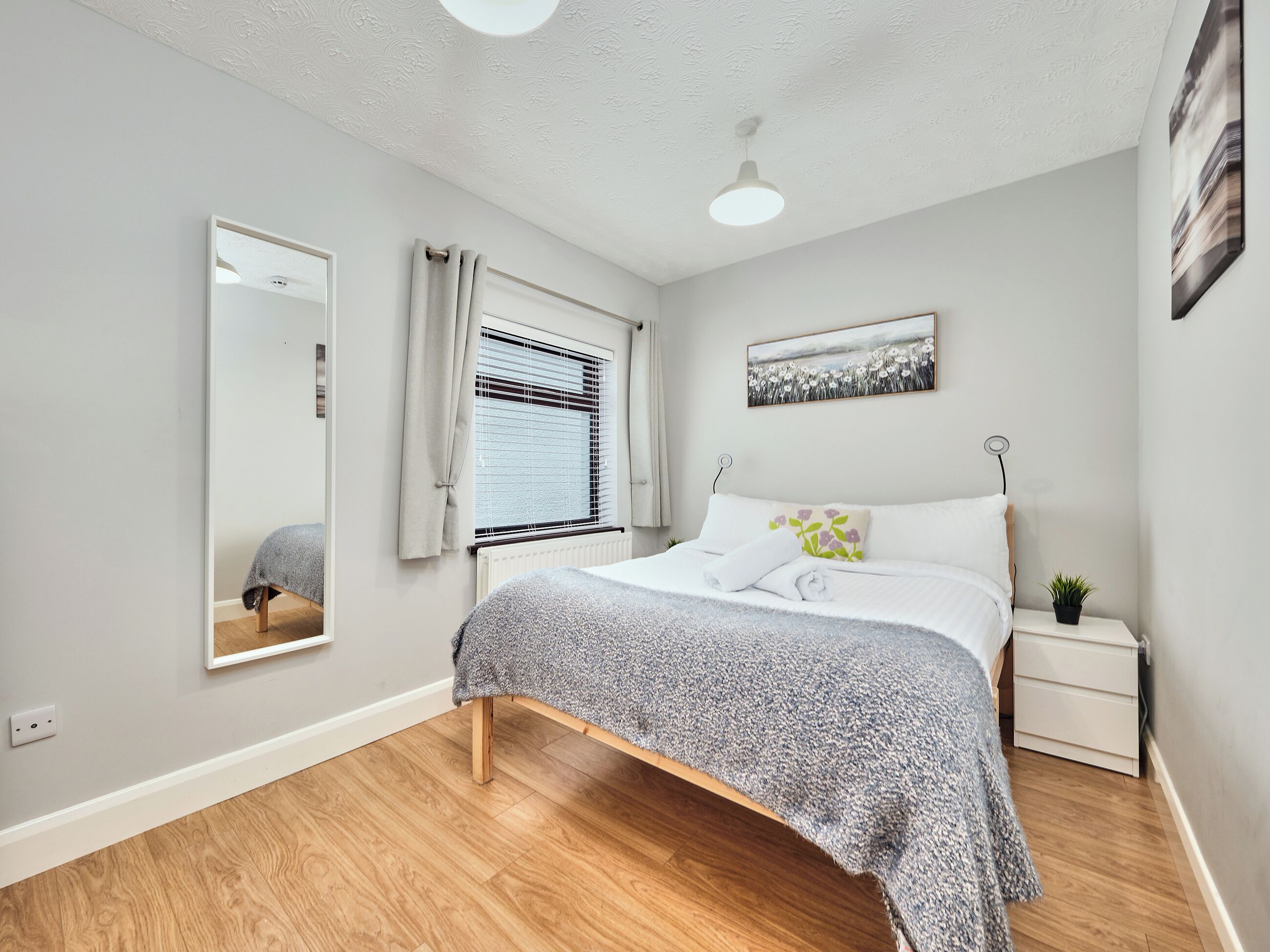 A bedroom with a bed, a bedside table with a plant, a window with curtains, a ceiling light, and a wall-mounted picture.
