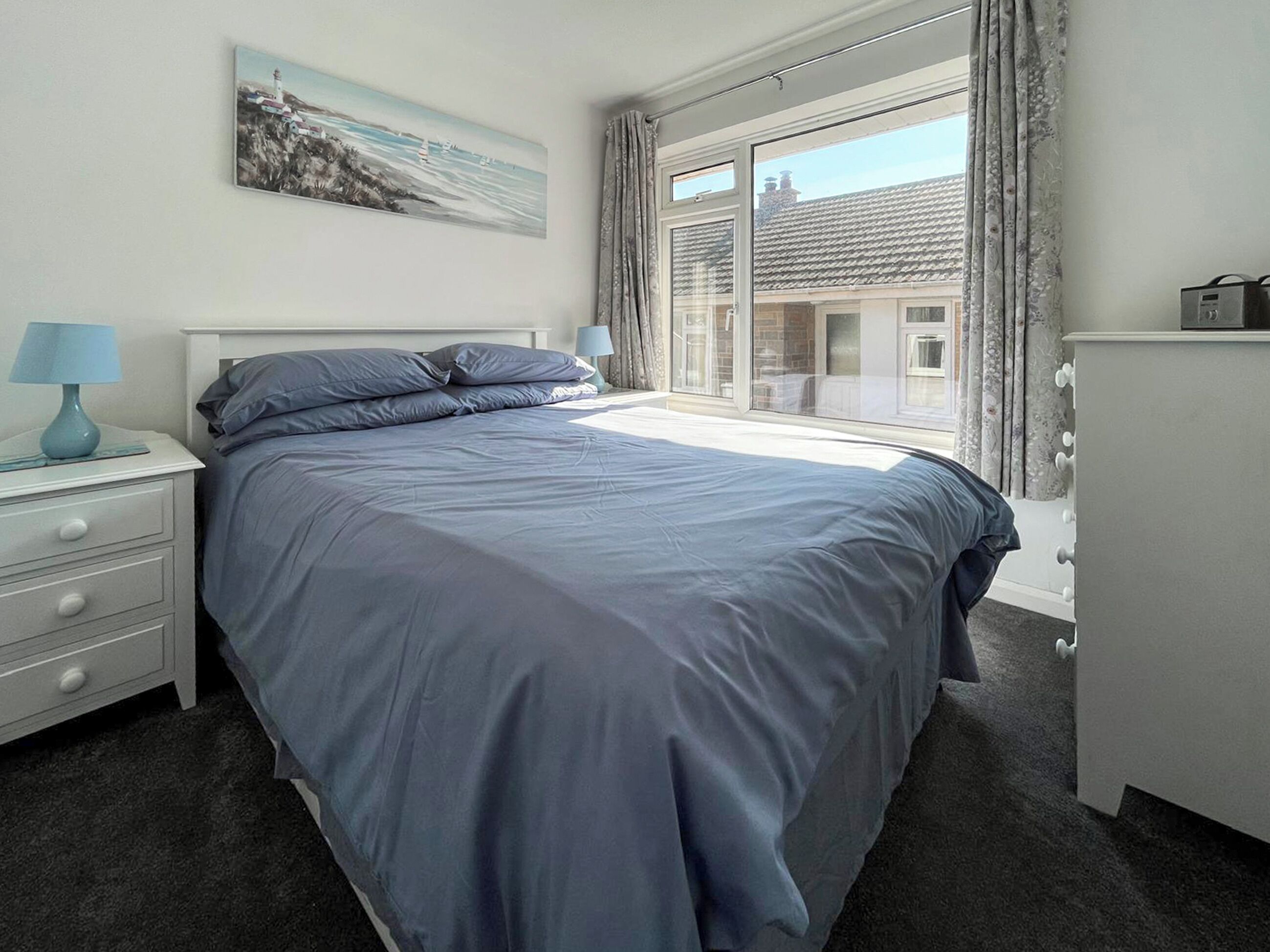 A bedroom with a bed, a nightstand, a dresser, a window with curtains, and a painting on the wall.