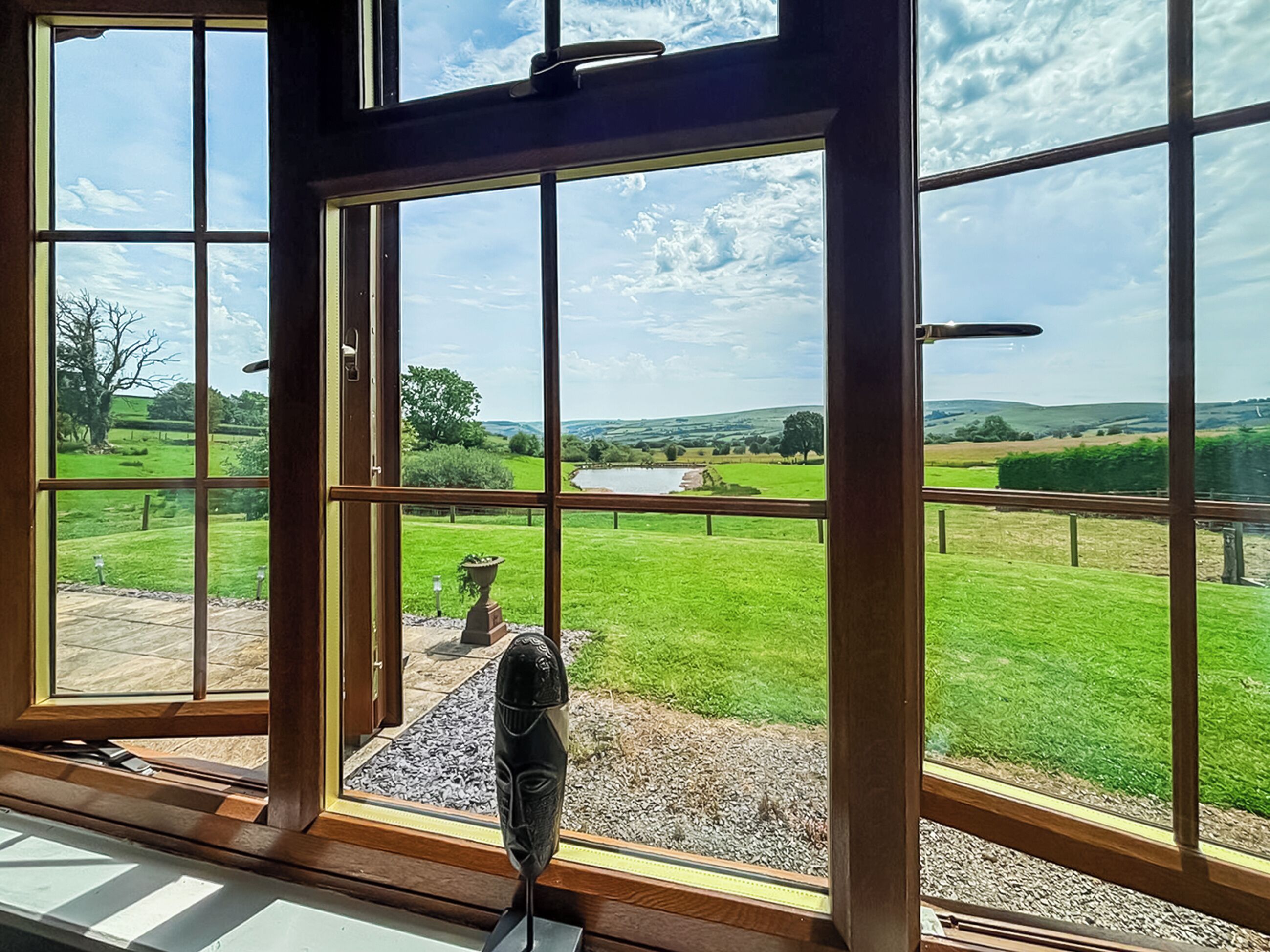 Ein großes Fenster mit Blick auf eine grüne Landschaft, darunter ein Teich und sanfte Hügel.