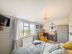 Quarto com cama, escrivaninha, cadeira, televisão e janela com cortinas.