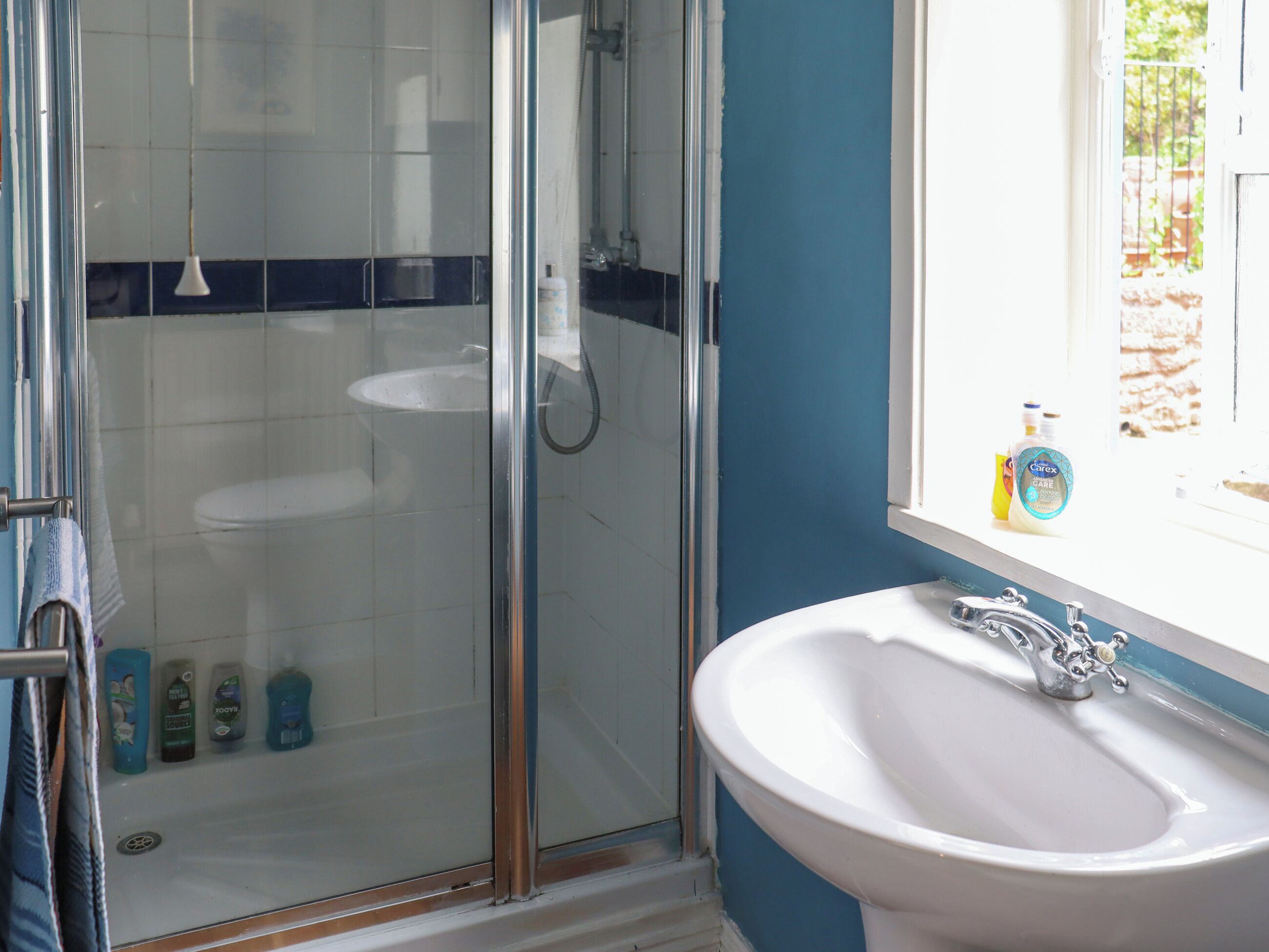A bathroom with a glass shower enclosure, a white sink, and a window with toiletries on the sill.
