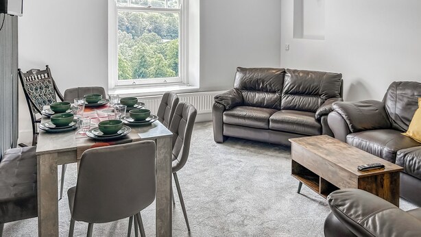 Een moderne woonkamer met een eethoek, leren banken, een houten salontafel en uitzicht op groen door een groot raam.