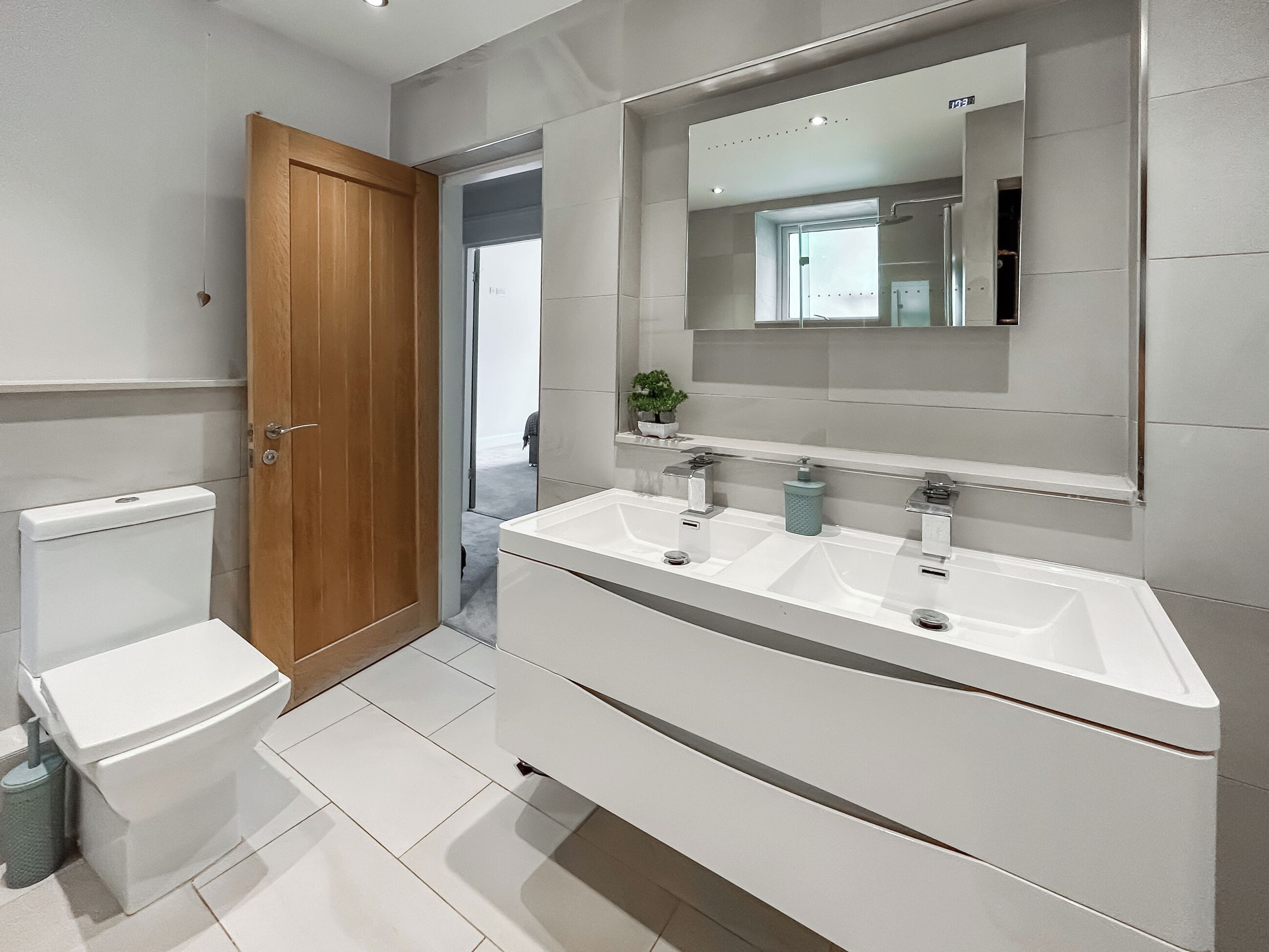 Une salle de bain moderne avec une porte en bois, des toilettes blanches et un grand miroir au-dessus d’un lavabo blanc.