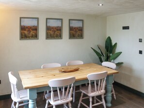 Een eetruimte met een houten tafel en witte stoelen, afbeeldingen aan de muur en een grote plant.