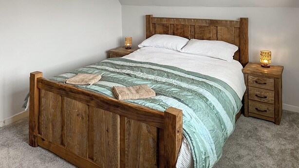 Ein hölzernes Bett mit einer grün-weißen gestreiften Tagesdecke, zwei Nachttische mit Lampen und ein Holz-Kopfstück.