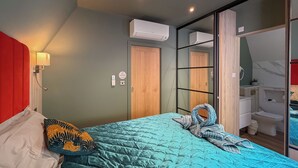 Quarto com cama, cabeceira vermelha, ar-condicionado fixado na parede, porta de madeira e banheiro visível por meio de um painel de vidro.