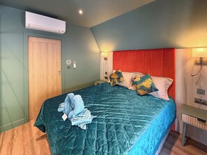 Ein Schlafzimmer mit einer türkisblauen Tagesdecke, zwei Kissen mit Blattmuster, einem roten Kopfteil, einer Holztür und einem Klimagerät.