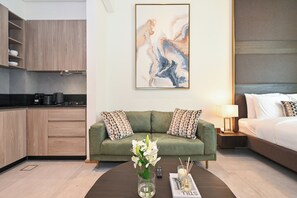 A modern hotel room with a green sofa, a coffee table with flowers, a painting on the wall, and a bed with white linens. - Bloomfields Studio in Signature Living South - JVC (Jumeirah Village Circle)