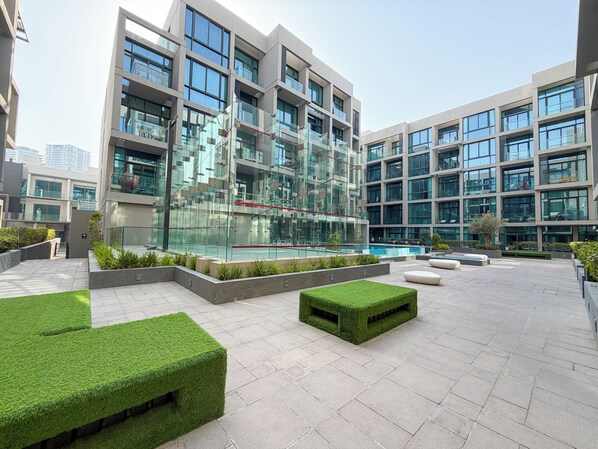 A modern building with multiple floors, large windows, and a courtyard with artificial grass and seating areas. - Bloomfields Studio in Signature Living South - JVC (Jumeirah Village Circle)