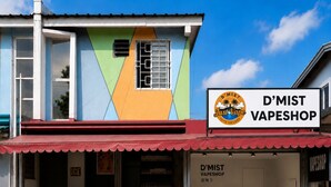 Un bâtiment à la façade colorée, avec une enseigne indiquant « D’MIST VAPESHOP » et une devanture de magasin pour « KBWT HOMESTAY ».