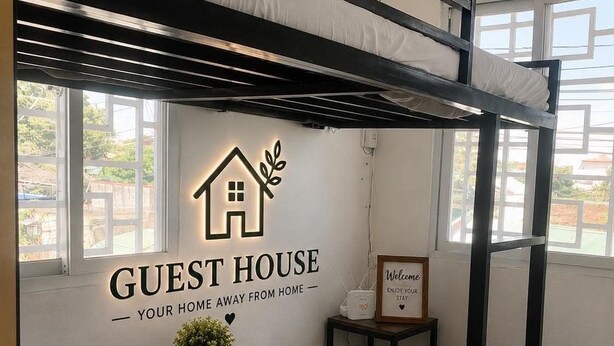 Une chambre avec des lits superposés, une table en bois et une petite plante en pot. Il y a une pancarte sur le mur indiquant « MAISON D’HÔTES » et une autre qui dit « BIENVENUE, PASSEZ UN BON SÉJOUR ».