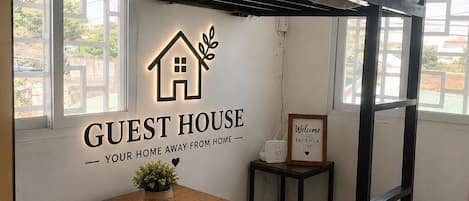 A bunk bed room with a wooden table and a small potted plant. There is a sign on the wall that reads 'GUEST HOUSE' and another sign that says 'WELCOME ENJOY YOUR STAY'.