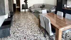 A modern living room with a grey sofa, a wooden dining table, and a kitchen area with a black cabinet. - Koras 142 - Guest House: A beautiful, bright apartment in the heart of Sennori (Sennori)