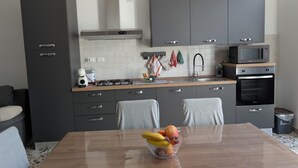 A modern kitchen with grey cabinets, a stainless steel range hood, a microwave, and an oven. There is a dining table with a bowl of fruit in the center. - Koras 142 - Guest House: A beautiful, bright apartment in the heart of Sennori (Sennori)