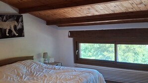 A bedroom with a large bed, a window with a view of trees, a wooden ceiling, and a painting on the wall. - Les Pierres Séches Vacation Rental, Les Andrieux 4/5 Banon (Banon)