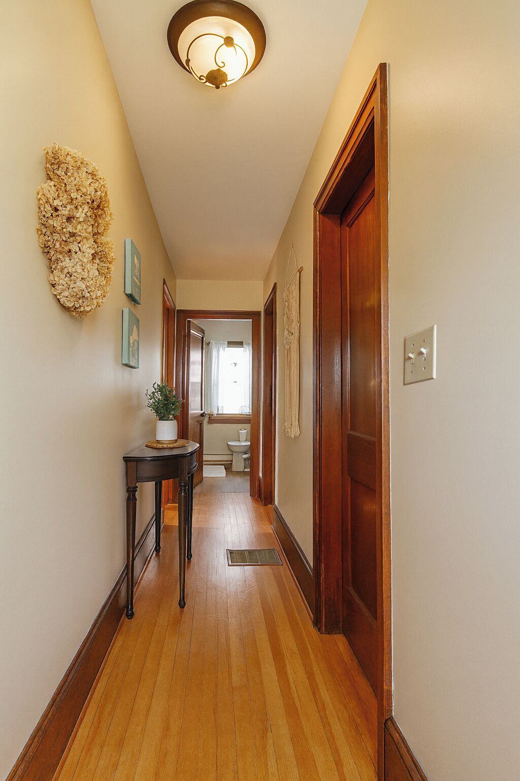 A hallway with wooden flooring, a wall-mounted light fixture, a small table with a plant, and a door leading to a bathroom.