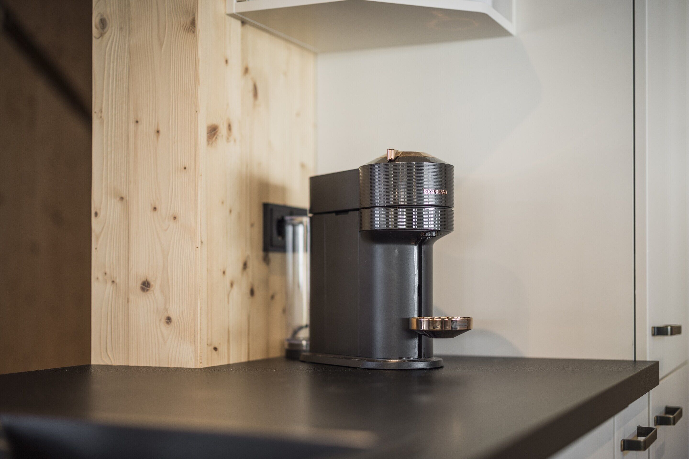 Eine Kaffeemaschine steht auf einer Arbeitsplatte, im Hintergrund sind ein Holzausschnitt und ein weißes Schranksystem zu sehen.