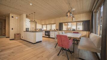 A spacious room with wooden floors and walls, a central dining table with chairs, a sofa, and a kitchen area with cabinets and appliances.