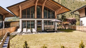 Ein modernes Haus mit großen Fenstern, einer Holzterrasse und Blick auf einen bewaldeten Berg.