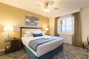 Habitación de hotel con cama, dos lámparas de noche, ventilador de techo, ventana con cortinas y vista al exterior.