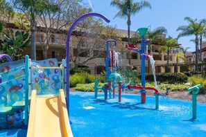 A children's play area with slides and water features, surrounded by palm trees and a building with balconies.