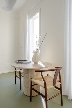 Ein Essbereich mit einem runden Tisch, zwei Stühlen, einer Vase mit Zweigen und einem Fenster mit Vorhängen.