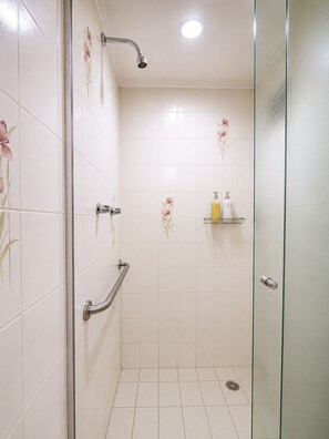 A glass-enclosed shower with white tiles and floral patterns, a handheld showerhead, a handheld shower control, and a small shelf with toiletries. - Fully equipped apartment in Campinas with pool, gym, and garage. (Campinas)